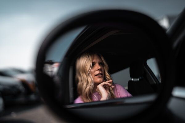 Die Spiegelung einer im Auto sitzenden Frau im Seitenspiegel, die fragend die Finger unter dem Kinn hält.